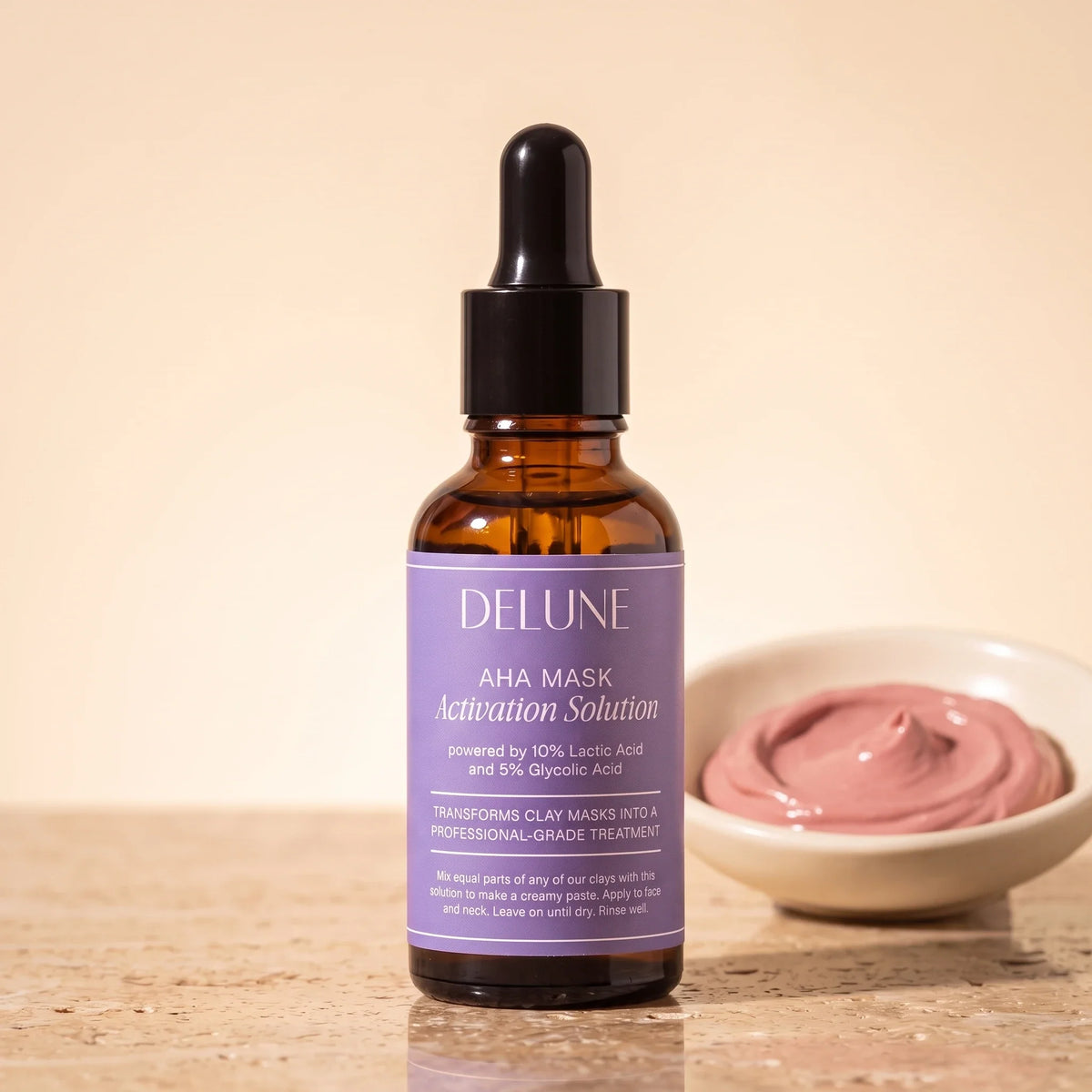 Delune AHA Mask Activation Solution 30ml dropper bottle on travertine beside a dish of rose clay paste