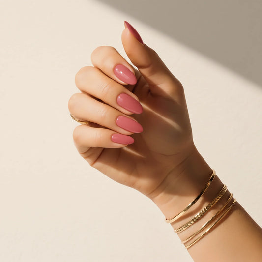 Hand wearing Delune Blush mauve-pink nail wraps with gold bangles against a warm plaster wall