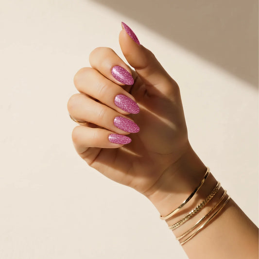 Hand wearing Delune Glitter Bug pink glitter nail wraps with gold bangles against a warm plaster wall