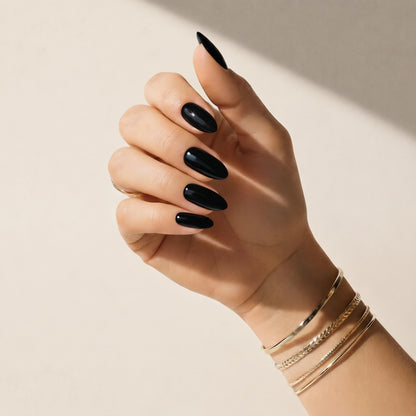 Hand wearing Delune Noir glossy black nail wraps with gold bangles against a warm plaster wall
