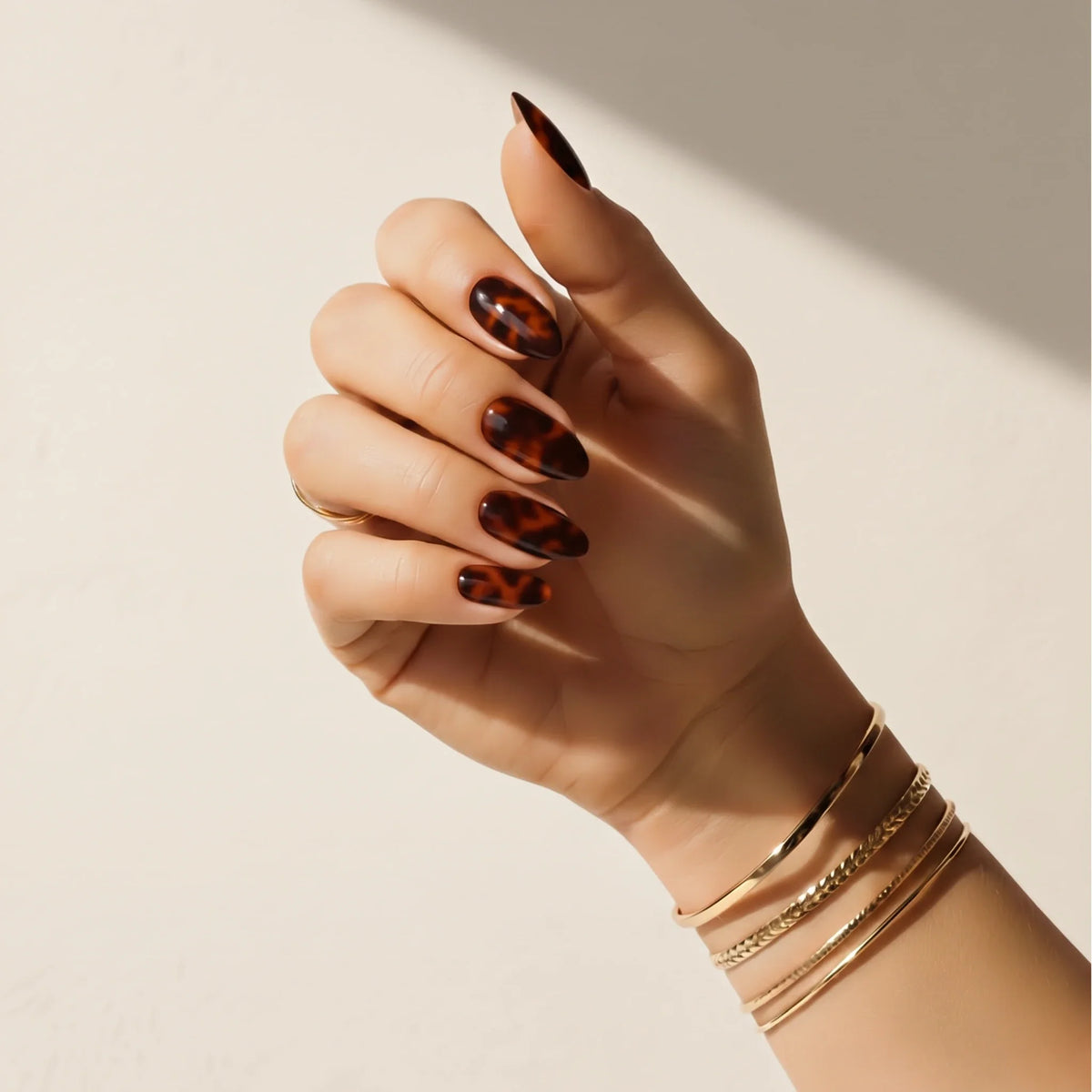Hand wearing Delune Tortoiseshell nail wraps with gold bangles against a warm plaster wall